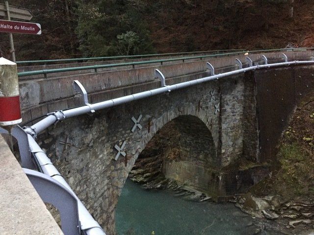 Pont en arc de pierre enjambant une rivière, avec garde-corps métalliques et panneau indicateur.