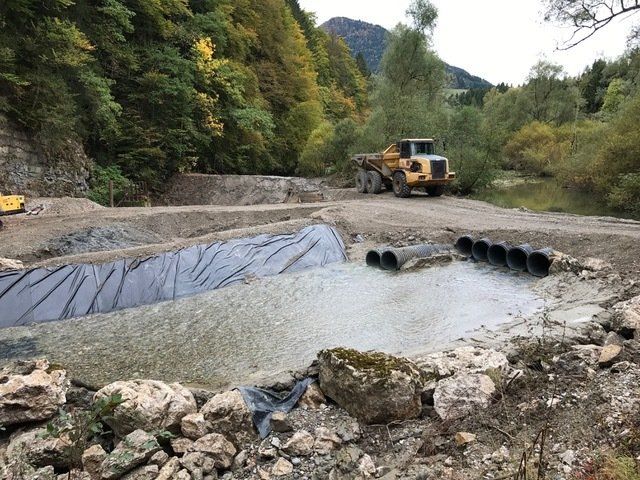 Chantier de construction avec engins lourds, terrassement et canalisations souterraines près d'une montagne et d'arbres.