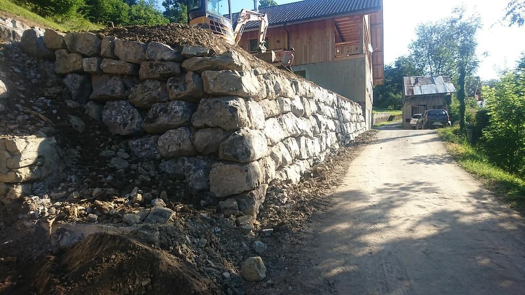 Mur de soutènement en pierre longeant un chemin de terre, avec un bâtiment en construction