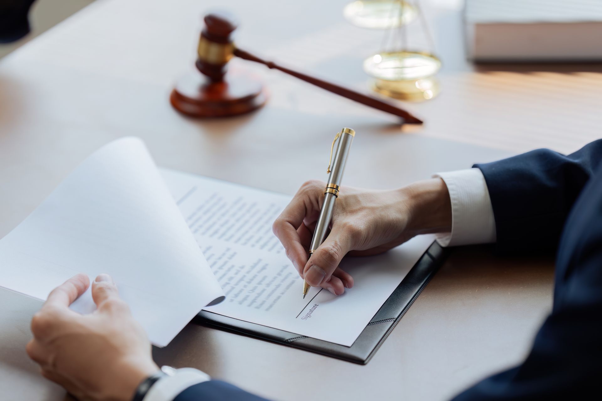 Une personne en costume sombre signe un document légal sur un bureau, à côté d'un maillet en bois et d'une balance de la justice.