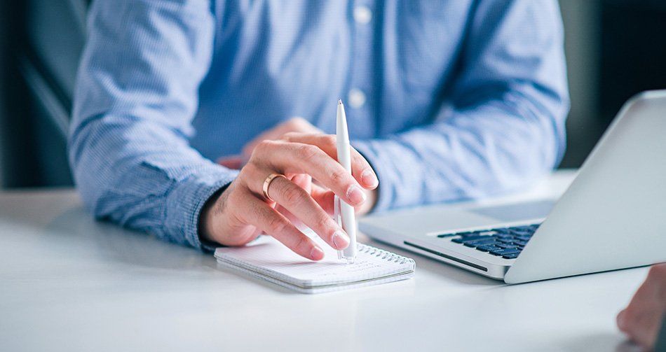Homme assis à un bureau devant un ordinateur portable, la main posée sur un bloc-notes