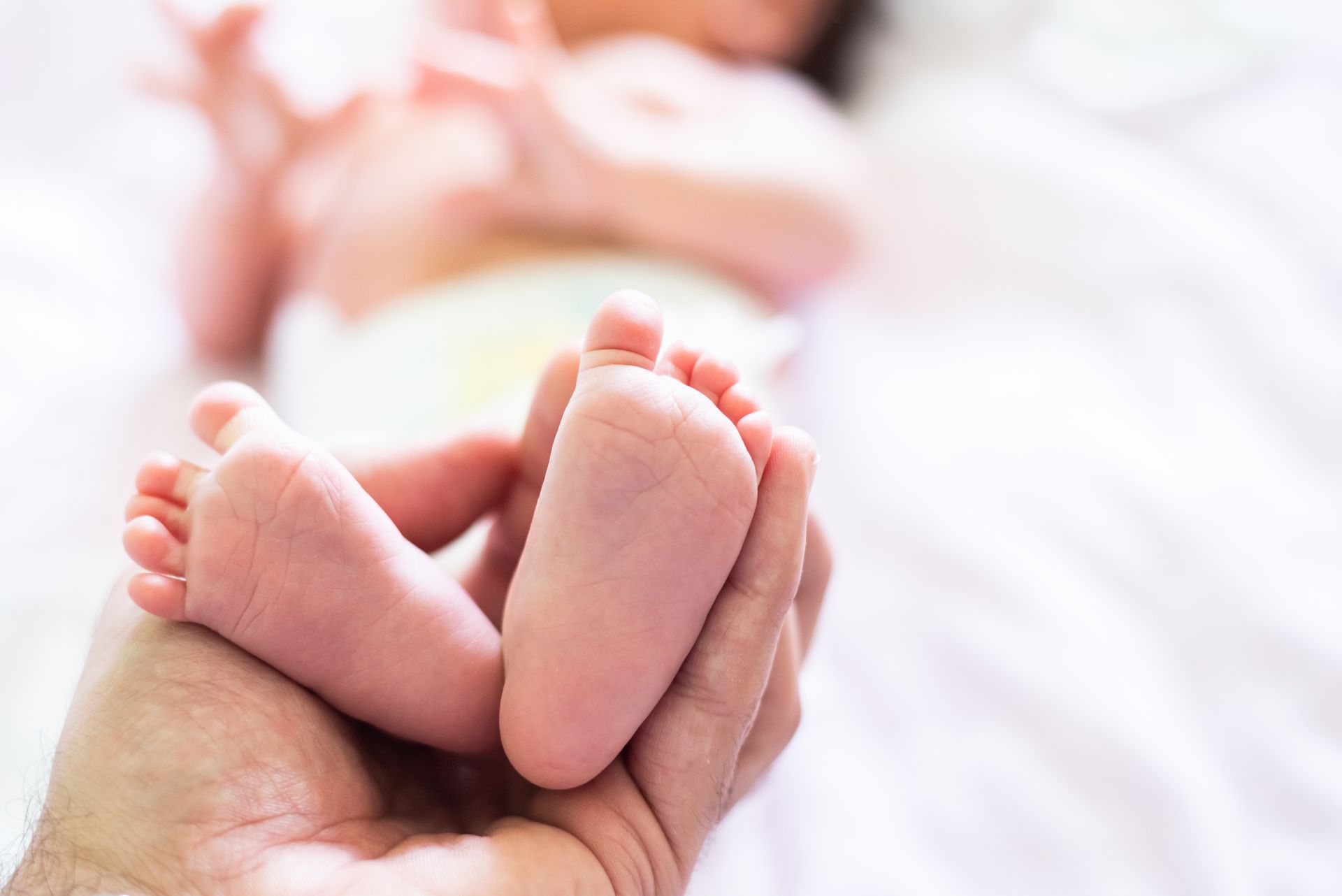 Une main berce les petits pieds d'un nouveau-né, sa peau rose et douce, le bébé est allongé sur un drap blanc.