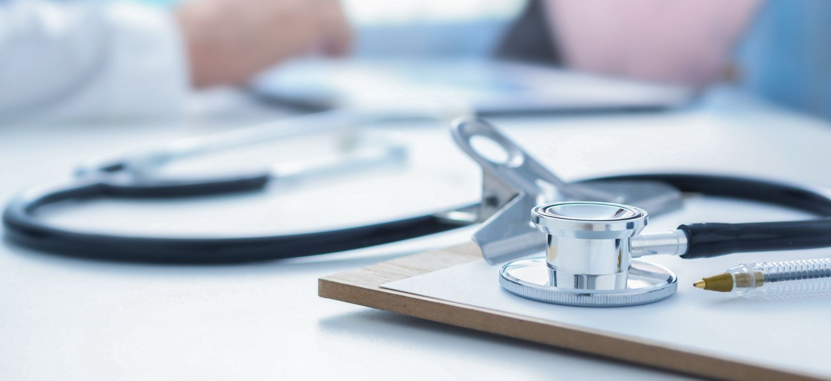 Un stéthoscope est posé sur un presse-papiers, avec un stylo. En arrière-plan, on aperçoit les mains d'un médecin.