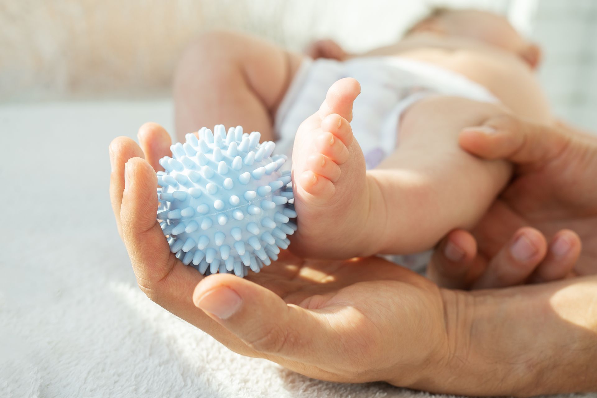 Le pied d'un bébé tenu avec une balle bleue à pointes dans la paume d'une main, sur une surface blanche.