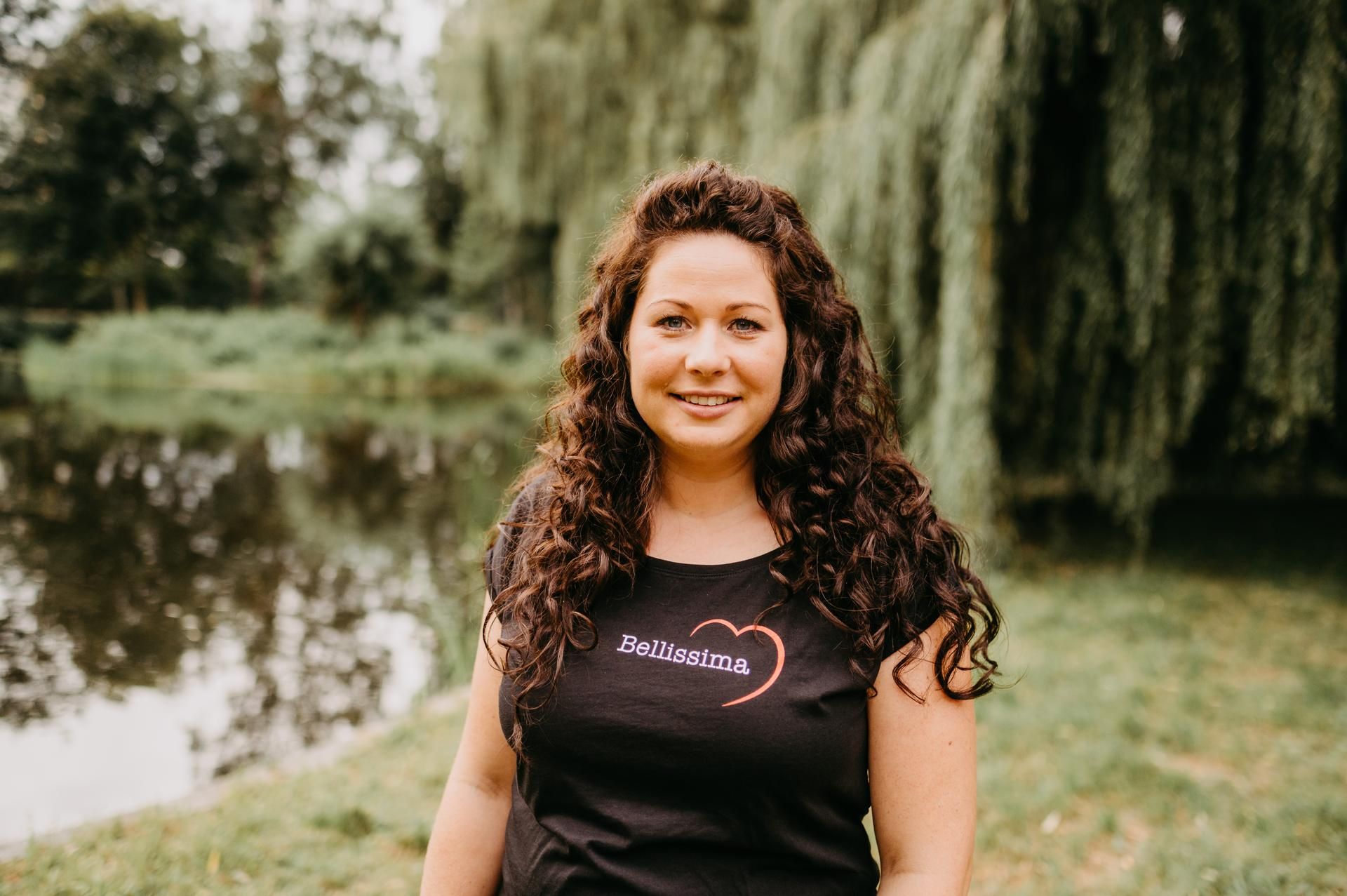 Eine Frau mit lockigem braunem Haar, die ein schwarzes T-Shirt trägt, lächelt vor einem Teich und Trauerweiden.