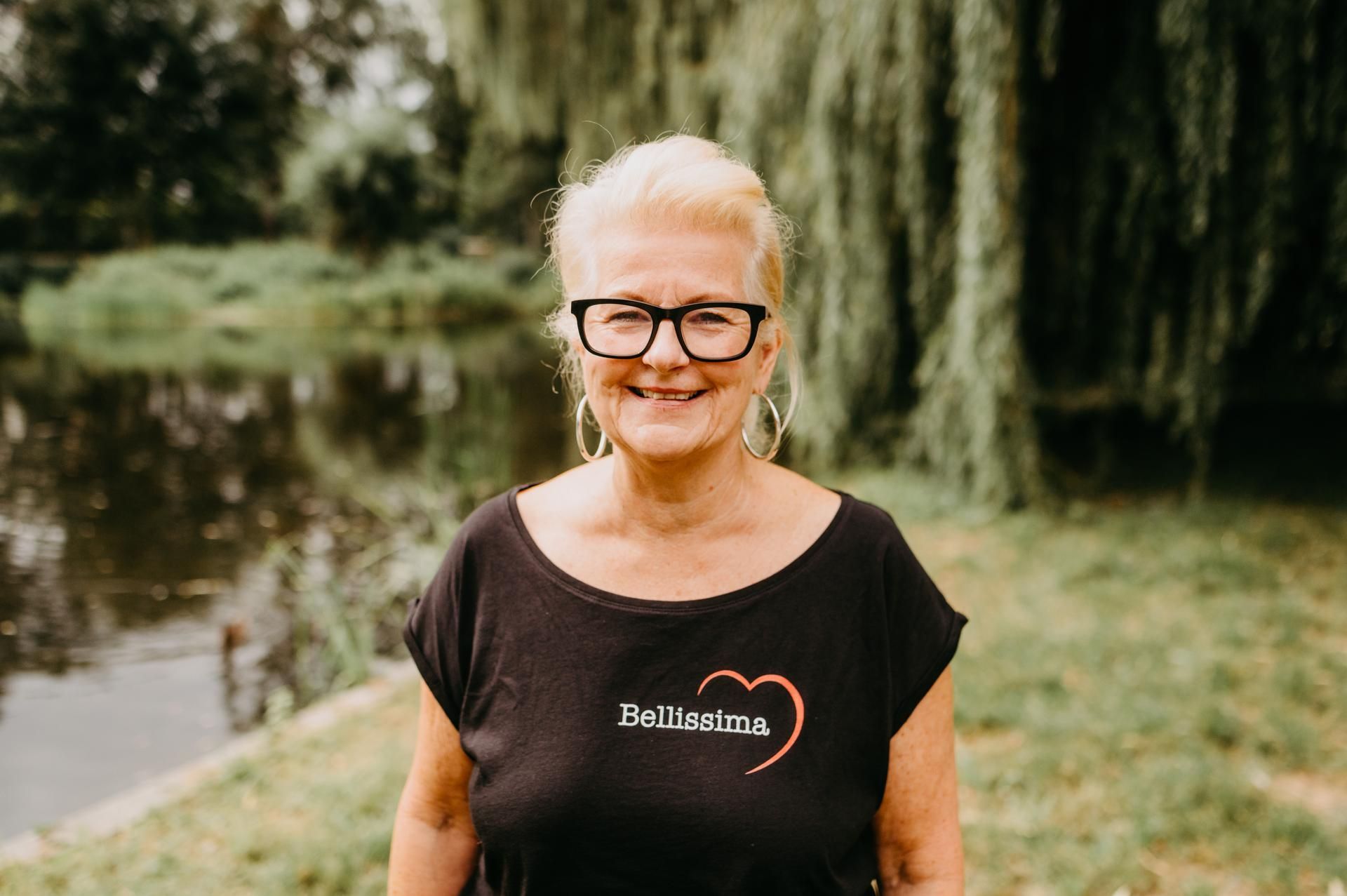 Eine Frau mit Brille und kurzen blonden Haaren, die ein schwarzes Hemd trägt, lächelt vor einem Gewässer mit Bäumen.