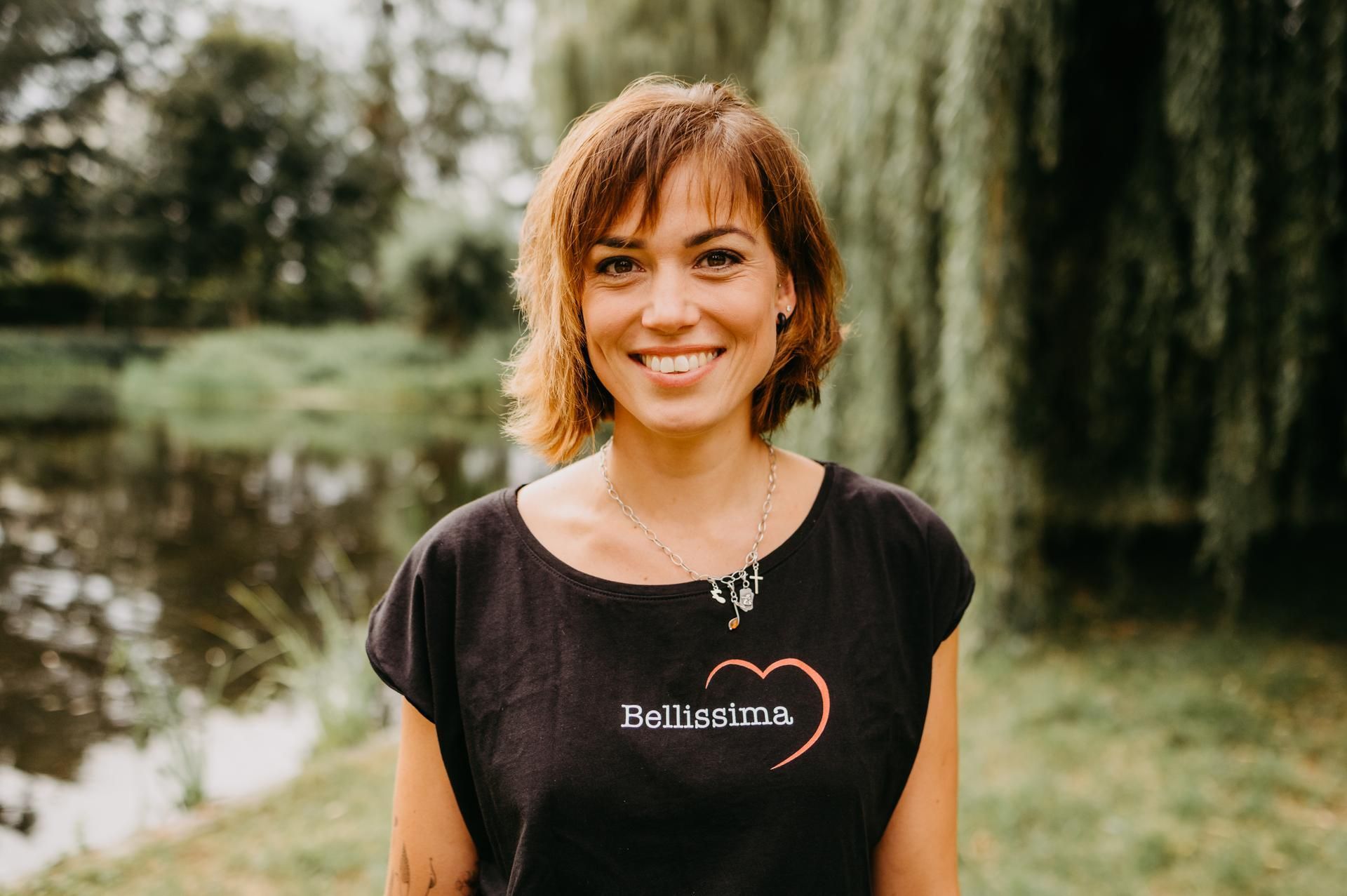 Eine Frau mit kurzen braunen Haaren lächelt. Sie trägt ein schwarzes T-Shirt mit Herz-Logo und steht im Freien in der Nähe eines Sees.