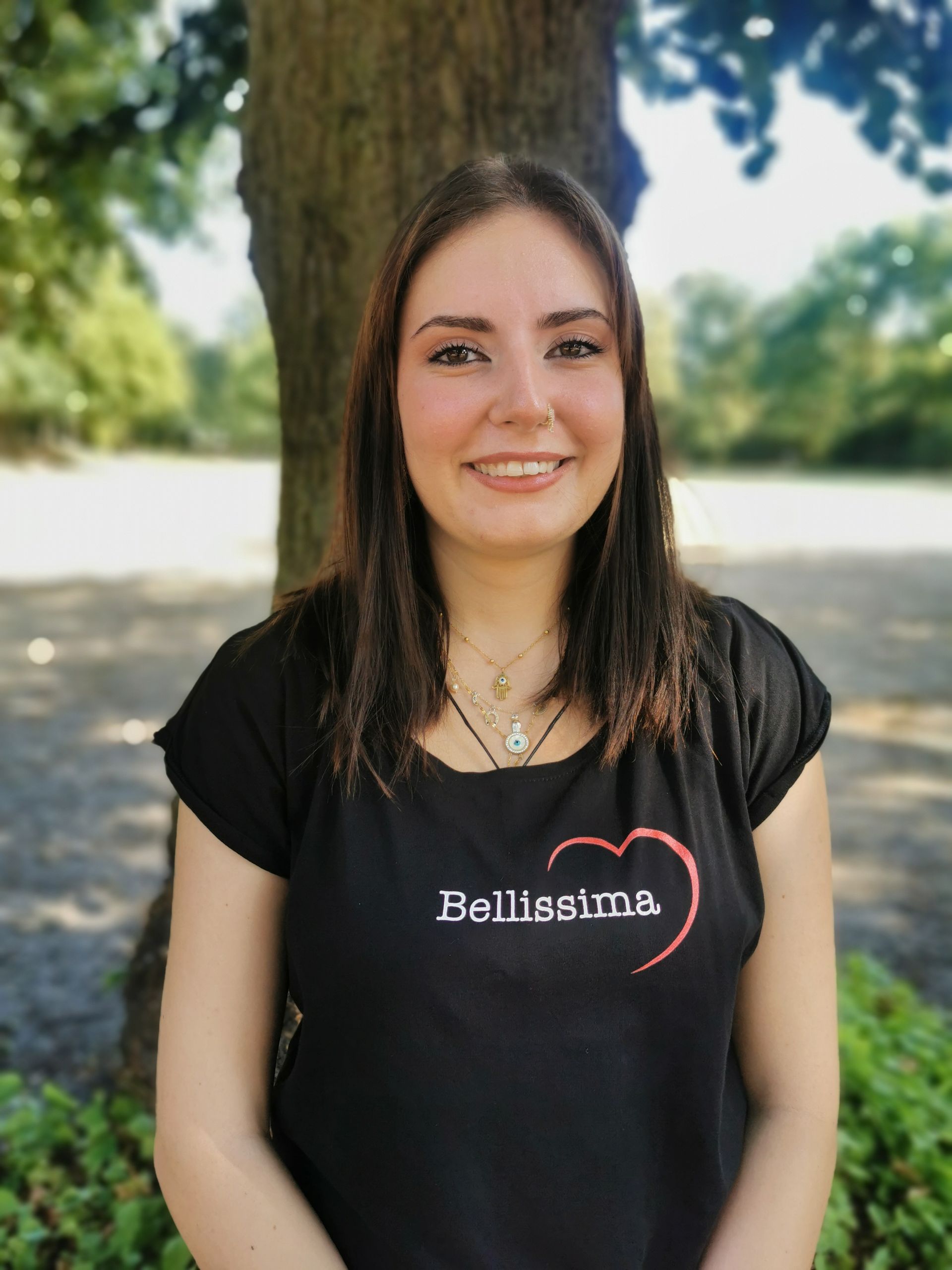 Eine Frau in einem schwarzen Hemd mit dem Aufdruck „Bellissima“ lächelt vor einem Baum und einer Parkkulisse.