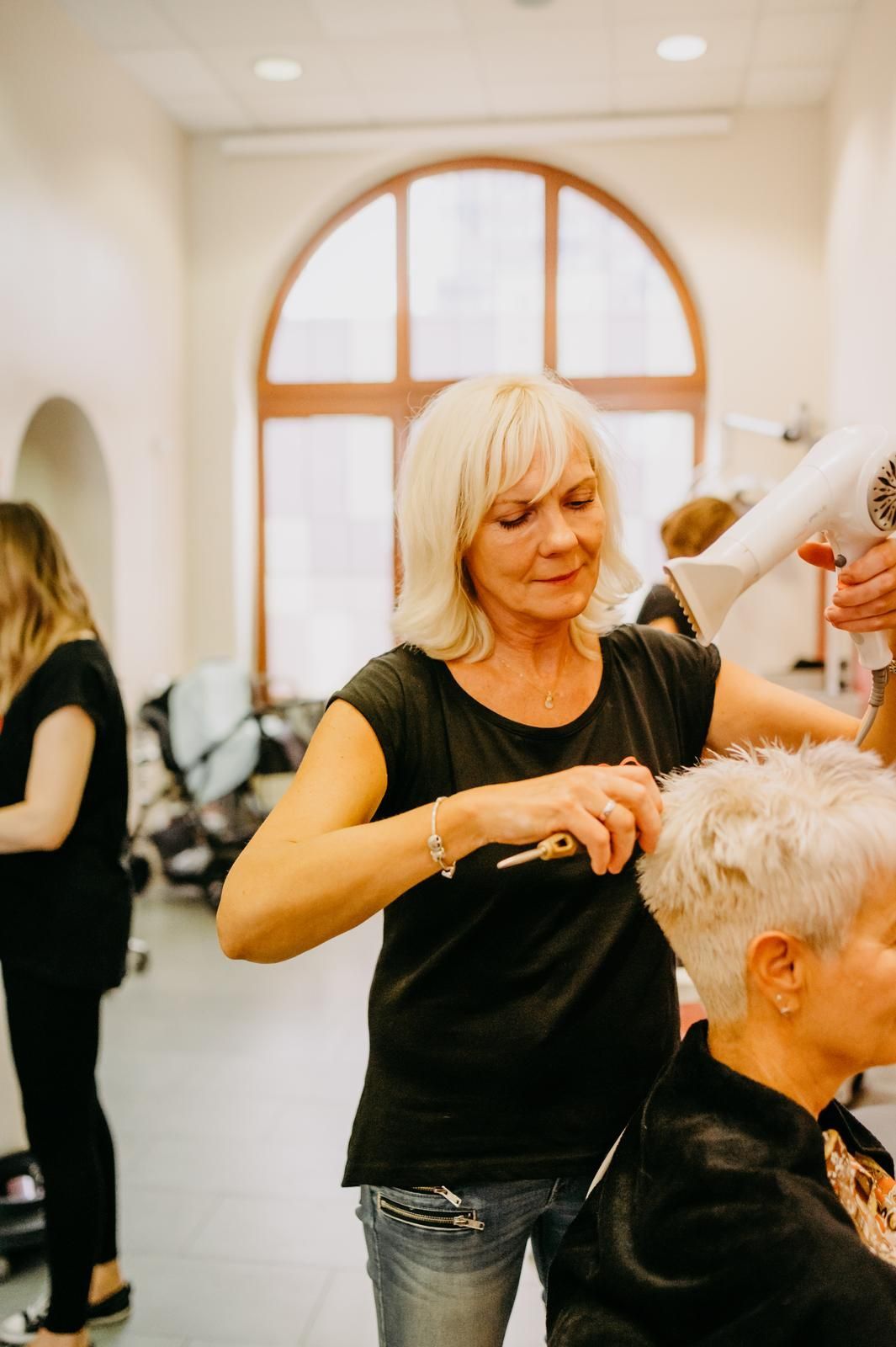 Eine Friseurin stylt im Salon die kurzen, grauen Haare einer Kundin mit Kamm und Föhn.