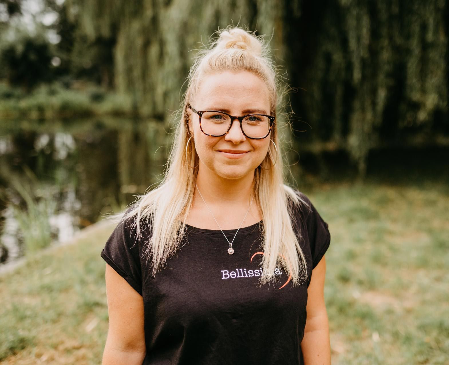 Eine Frau mit blonden Haaren und Brille in einem schwarzen Hemd steht draußen an einem Gewässer.