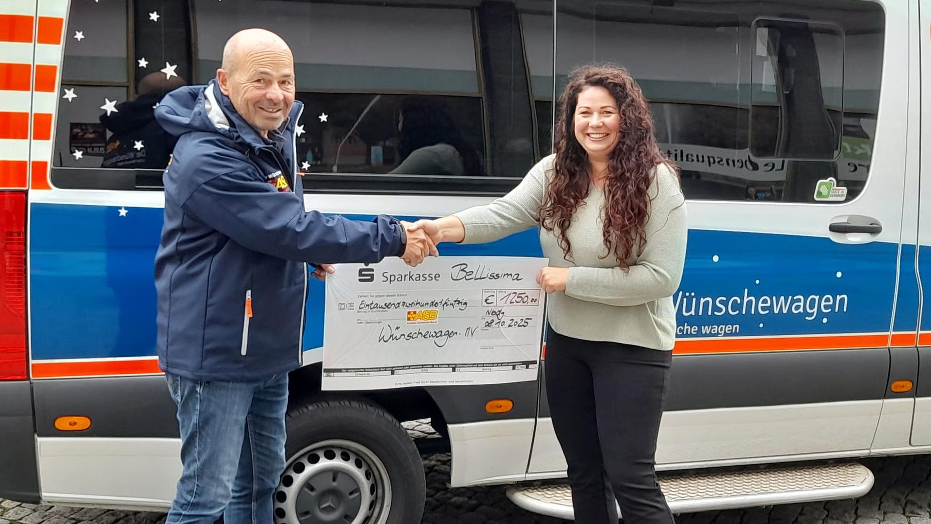Ein Mann überreicht einer Frau einen Scheck vor einem blau-weißen Lieferwagen mit der Aufschrift „Wünschewagen“.