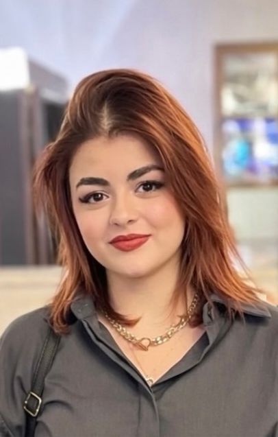 Frau mit roten Haaren und rotem Lippenstift, die ein graues Hemd und eine goldene Halskette trägt, lächelt. Verschwommener Hintergrund.