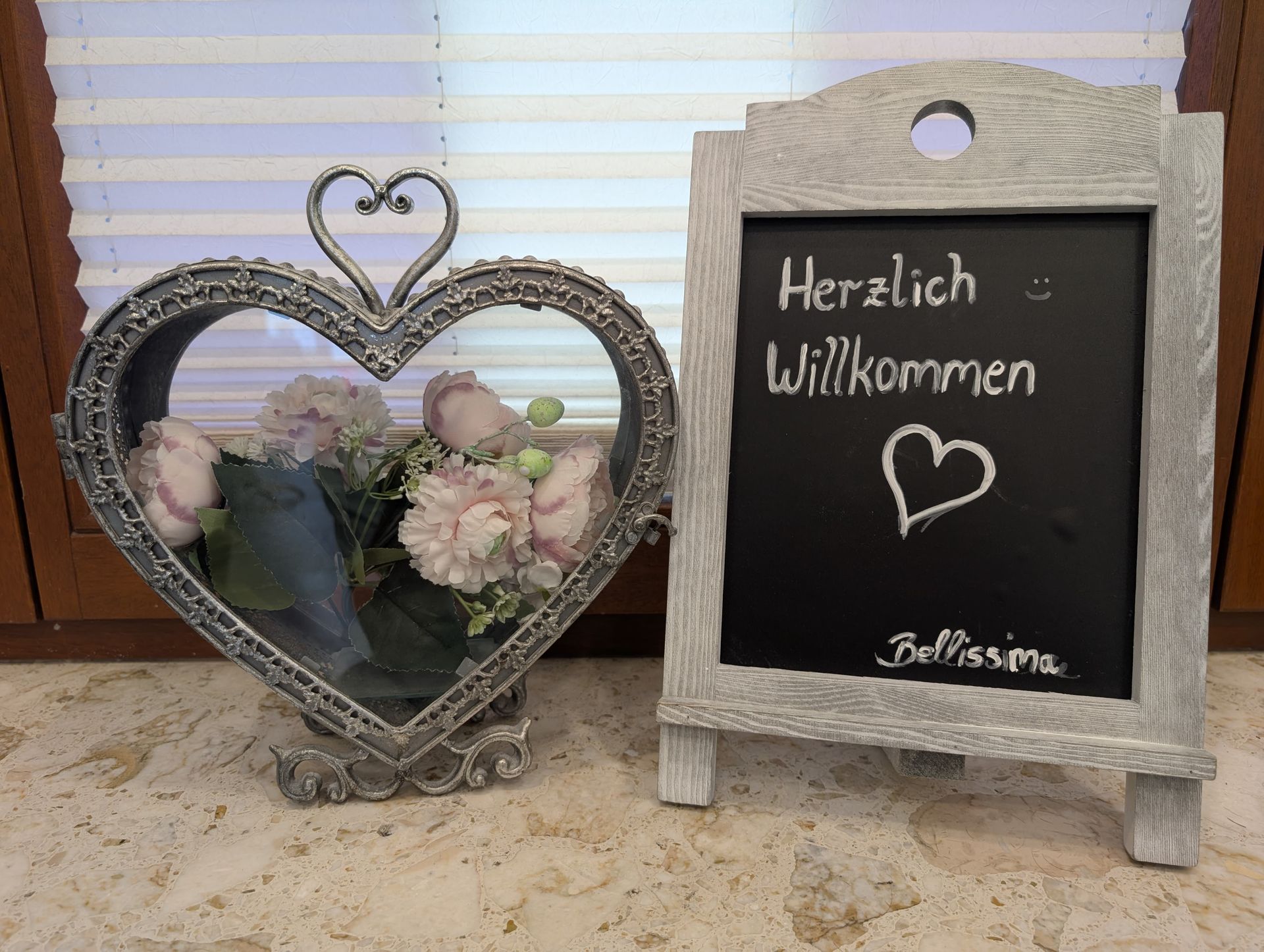 Herzförmige Dekoration mit Blumen neben einer Kreidetafel mit der Aufschrift „Herzlich Willkommen“.