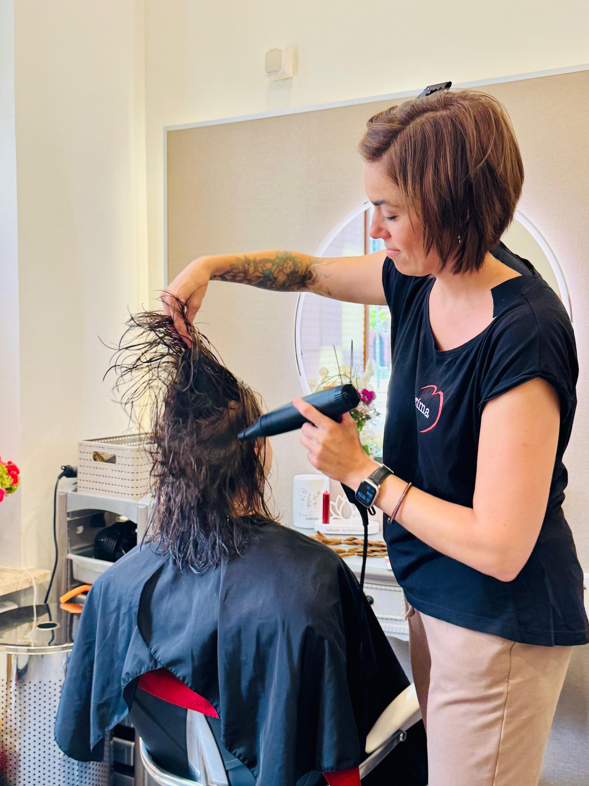 Eine Friseurin föhnt einer Kundin im Salon die nassen Haare. Die Friseurin trägt ein schwarzes Hemd und eine beige Hose.