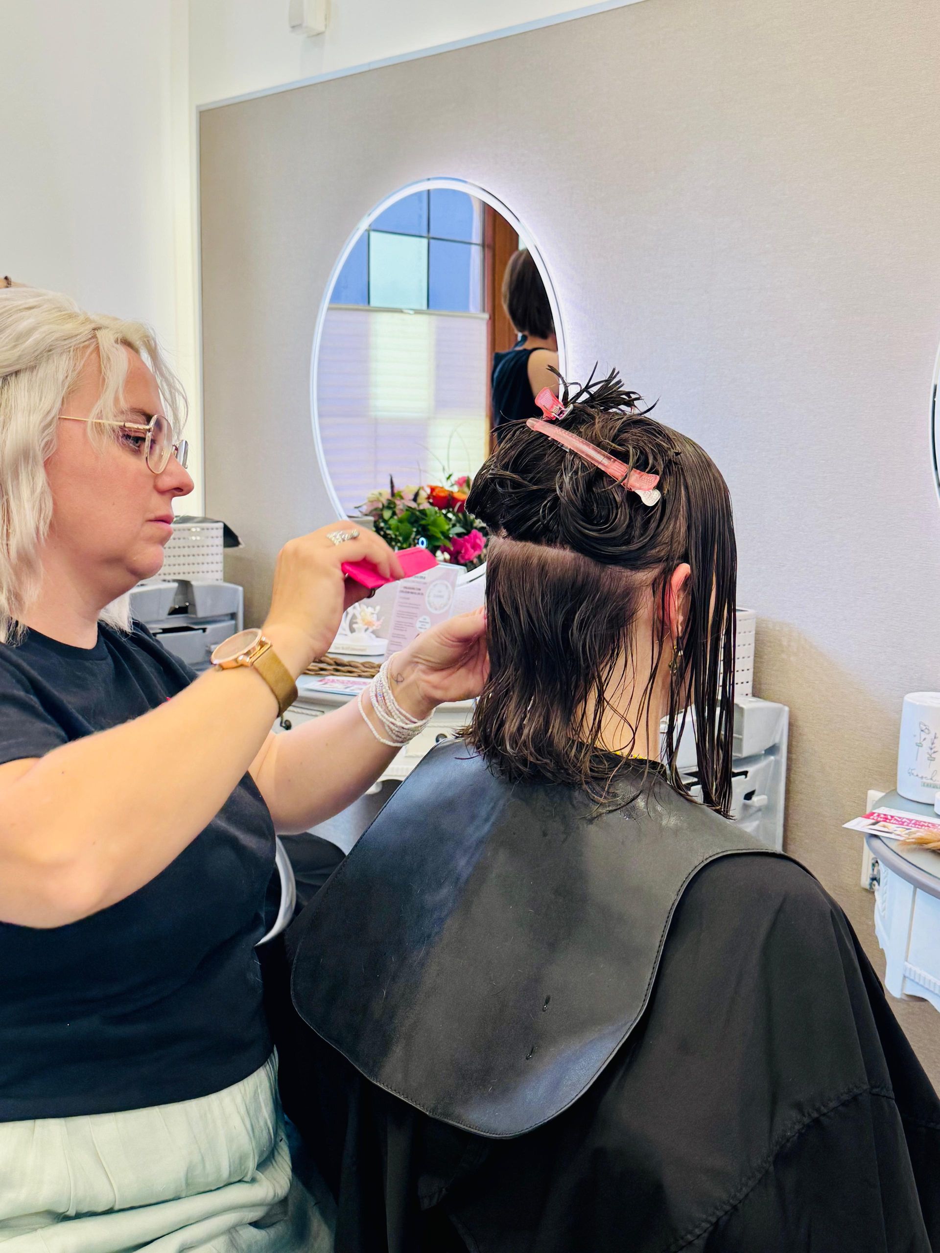 Eine Friseurin schneidet einer Kundin im Salon mit einer Haarschneidemaschine die nassen Haare. Die Kundin trägt einen schwarzen Umhang und ist abgewandt.