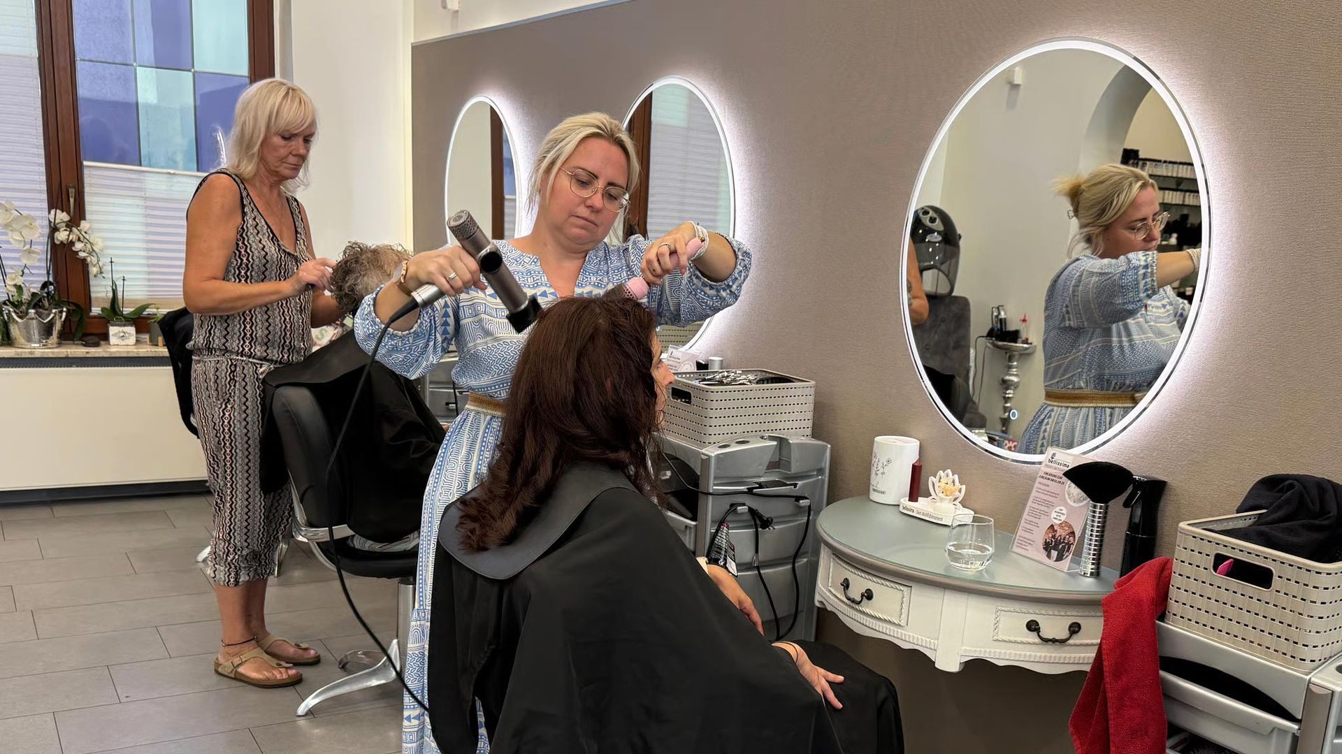 Friseure arbeiten in einem Salon an Kunden. Einer benutzt einen Föhn, die anderen schauen vor Spiegeln zu.