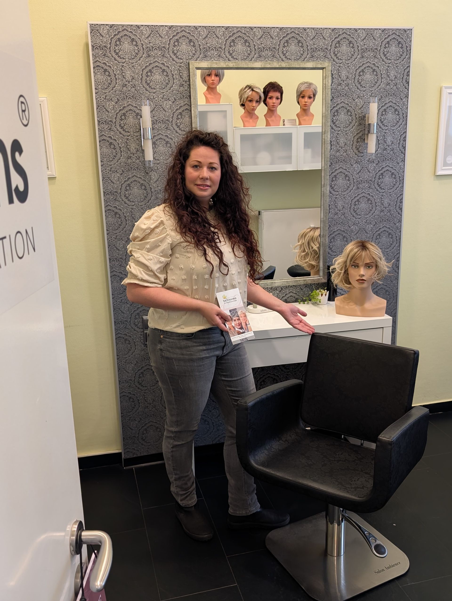 Frau in einem Stand mit Perückenauslage, die auf einen schwarzen Friseurstuhl zeigt.