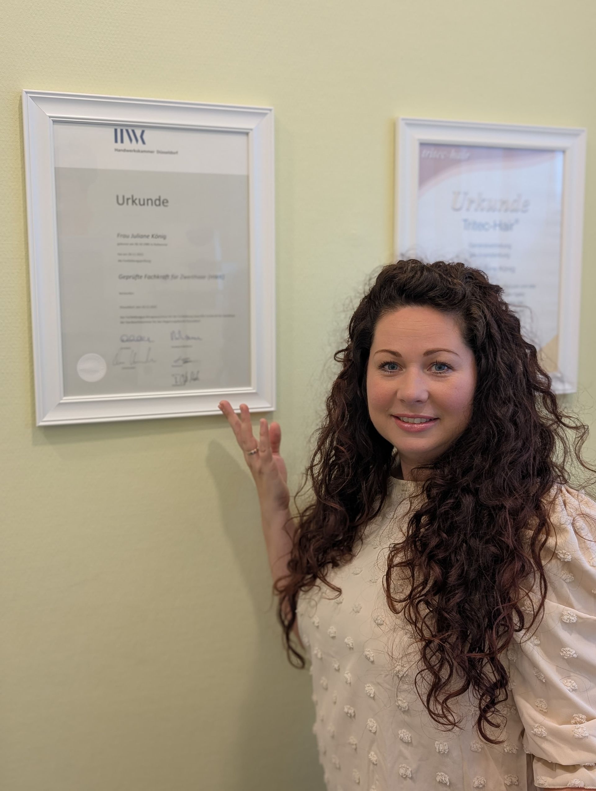 Eine Frau zeigt auf ein gerahmtes Zertifikat an einer gelben Wand; sie hat lockiges braunes Haar und trägt ein weißes, strukturiertes Oberteil.