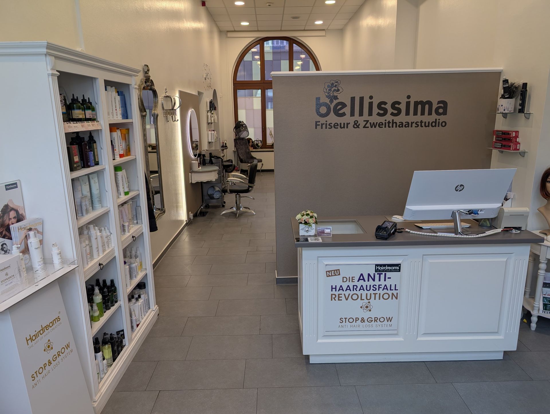 Interieur eines Friseursalons. Weißer Empfangstresen mit dem „bellissima“-Logo. Regale mit Haarpflegeprodukten. Stühle und Spiegel im Hintergrund.