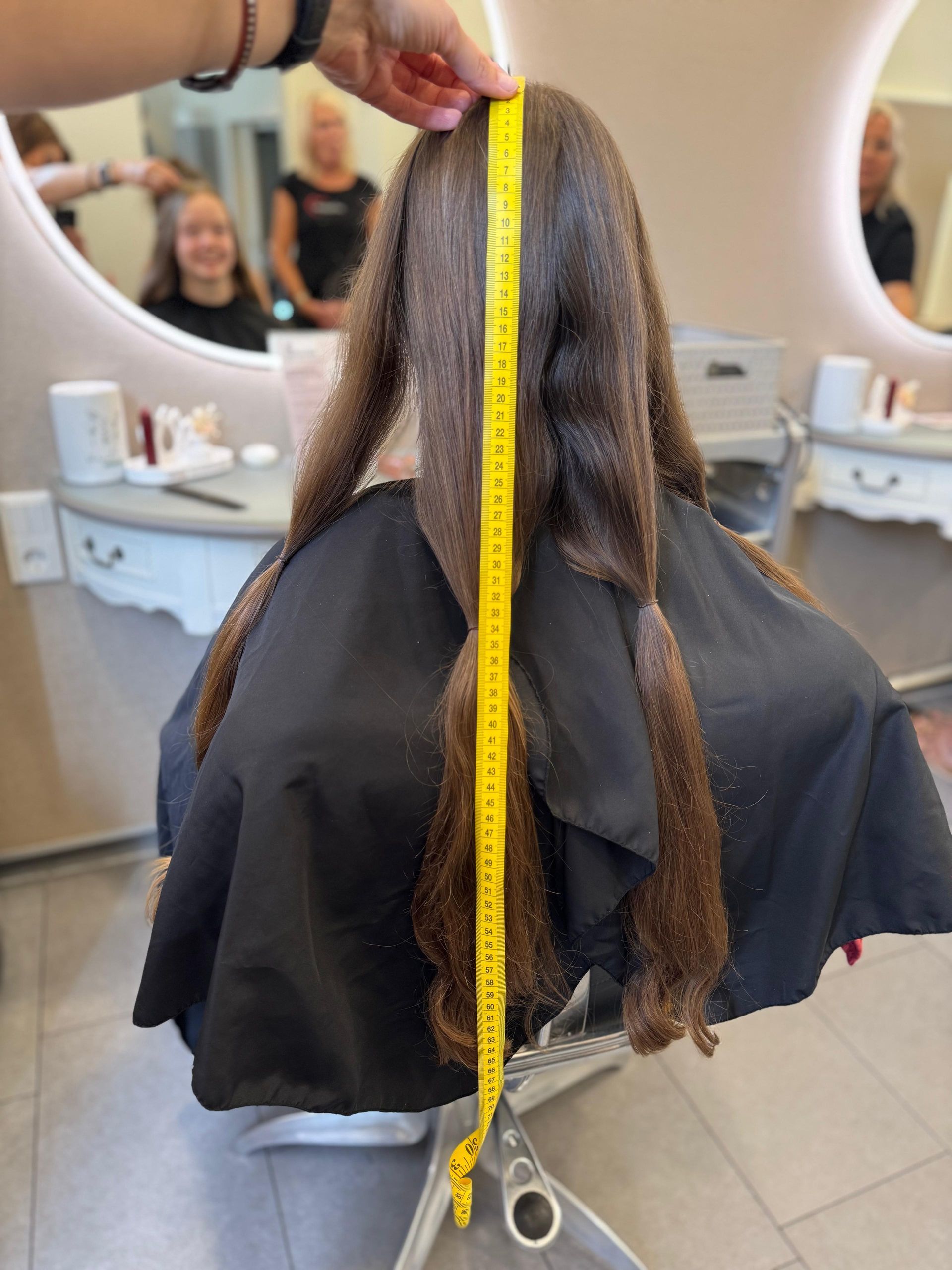 Die langen braunen Haare einer Person werden in einem Friseursalon mit einem gelben Maßband gemessen. Im Hintergrund sind weitere Personen zu sehen.