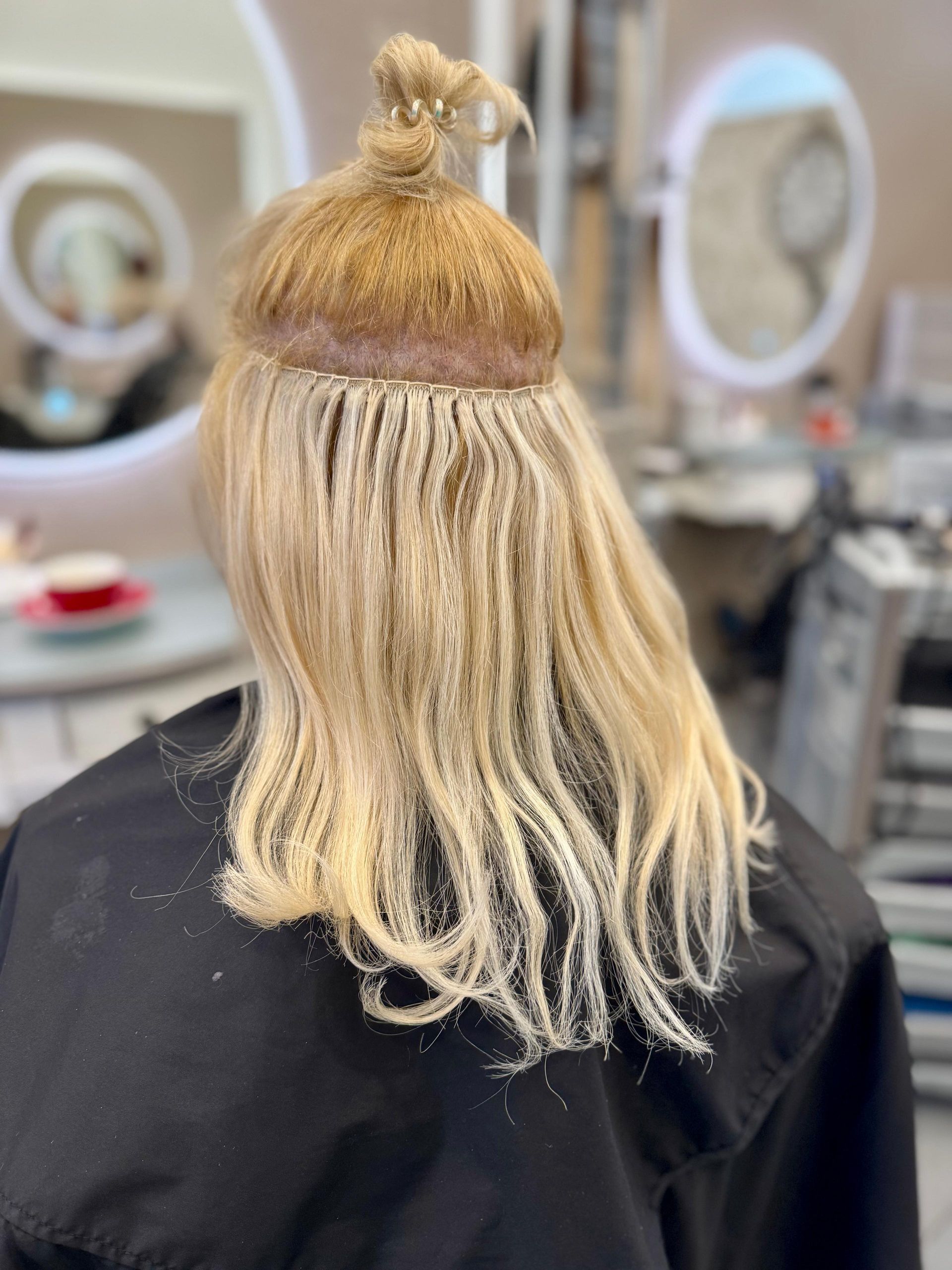 Frau mit blonden Haaren, mit dem Rücken zur Kamera, lässt sich in einem Salon Haarverlängerungen machen.