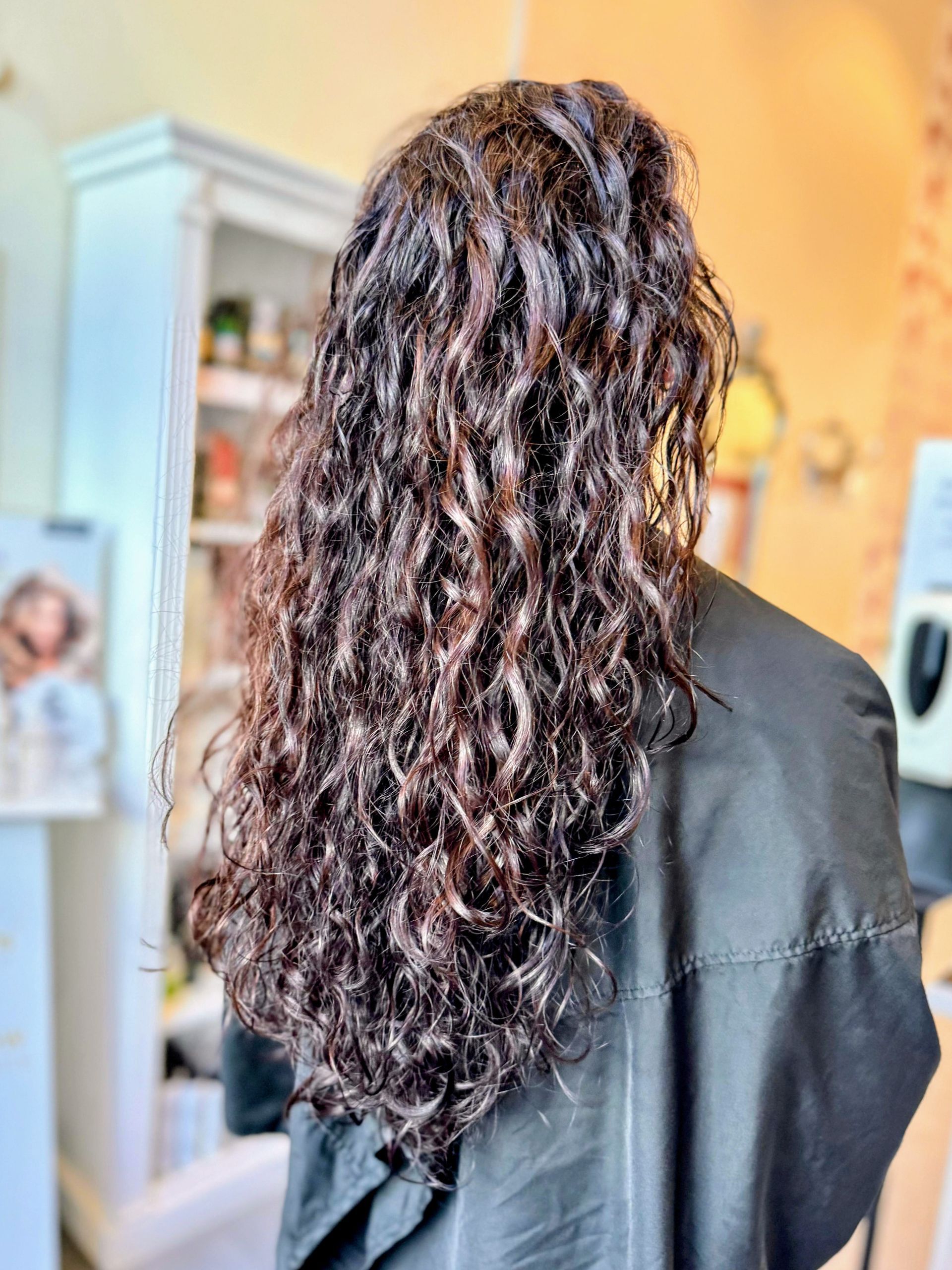 Langes, dunkles, lockiges Haar nach einem Friseurbesuch. Die Person trägt einen schwarzen Umhang; im Hintergrund sind ein weißer Schrank und eine weiße Wand zu sehen.