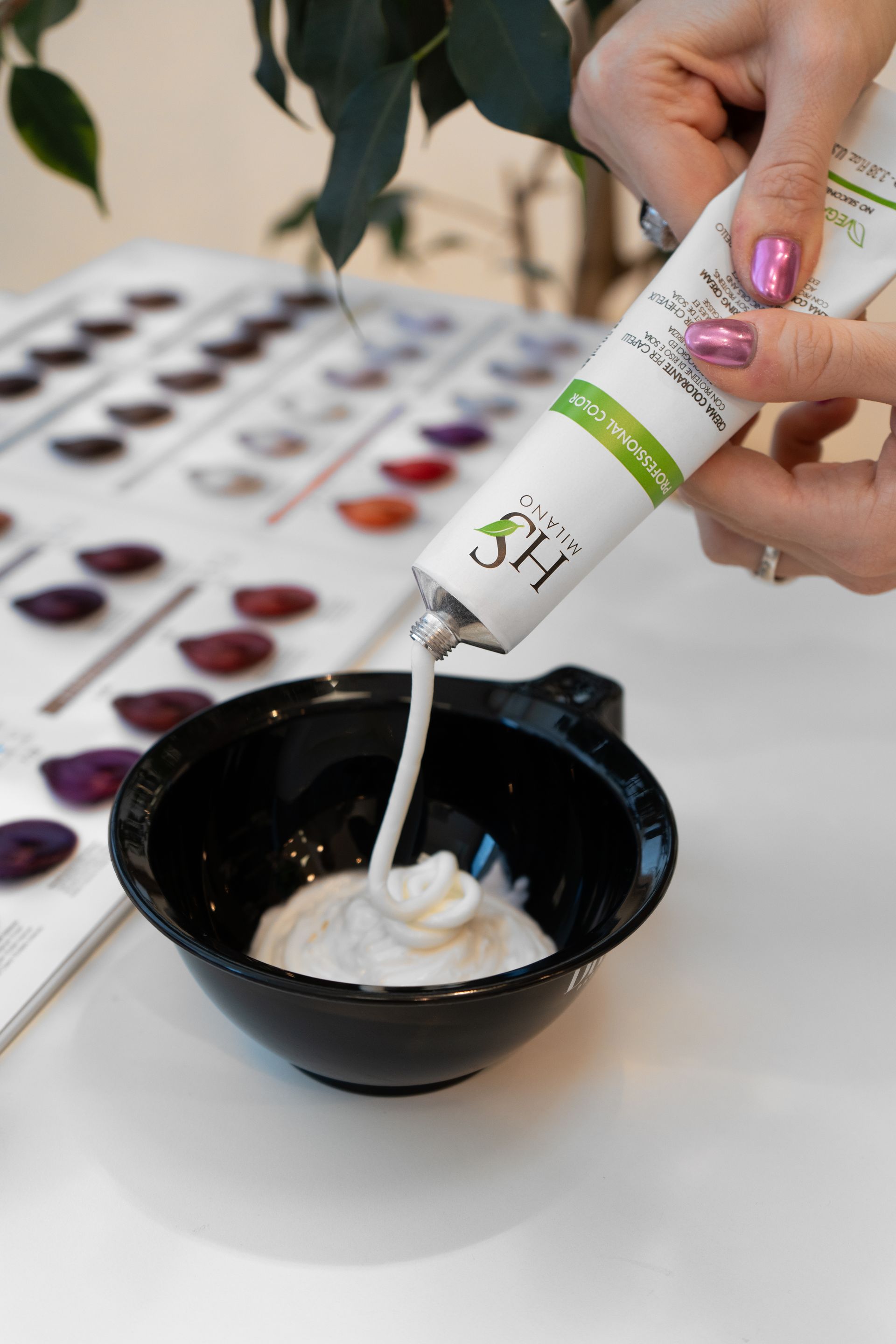 Eine Person drückt weiße Haarfarbe aus einer Tube in eine schwarze Schüssel; im Hintergrund sind Farbmuster zu sehen.