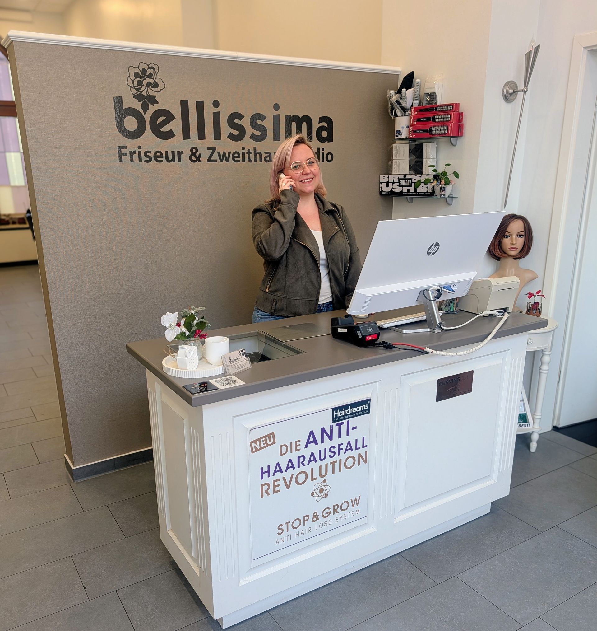 Eine Frau telefoniert an der Rezeption eines Friseursalons. Auf dem Empfangstresen stehen ein Computer und ein Schaufensterpuppenkopf.