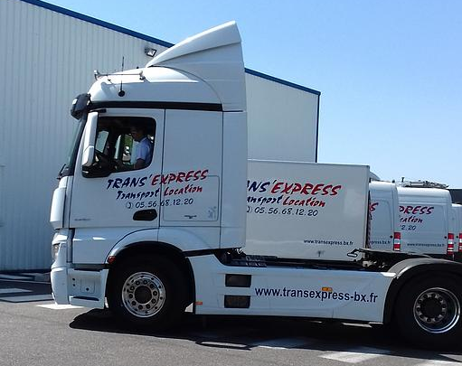 Semi-remorque Trans Express blanche garée devant un bâtiment. Conducteur visible. Ciel bleu, journée ensoleillée.