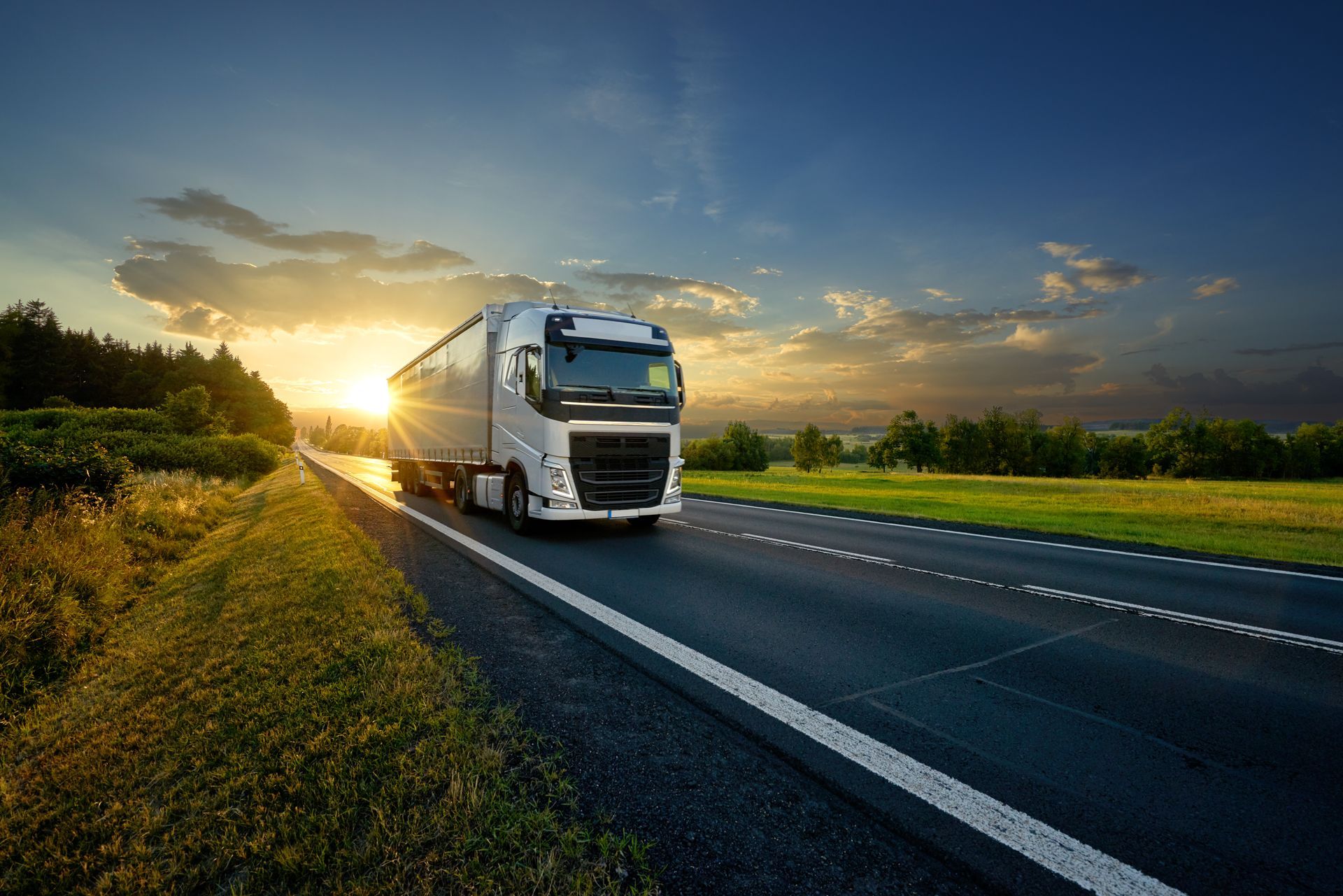 Semi-camion roulant sur une autoroute au coucher du soleil, avec un ciel bleu et des champs verts.
