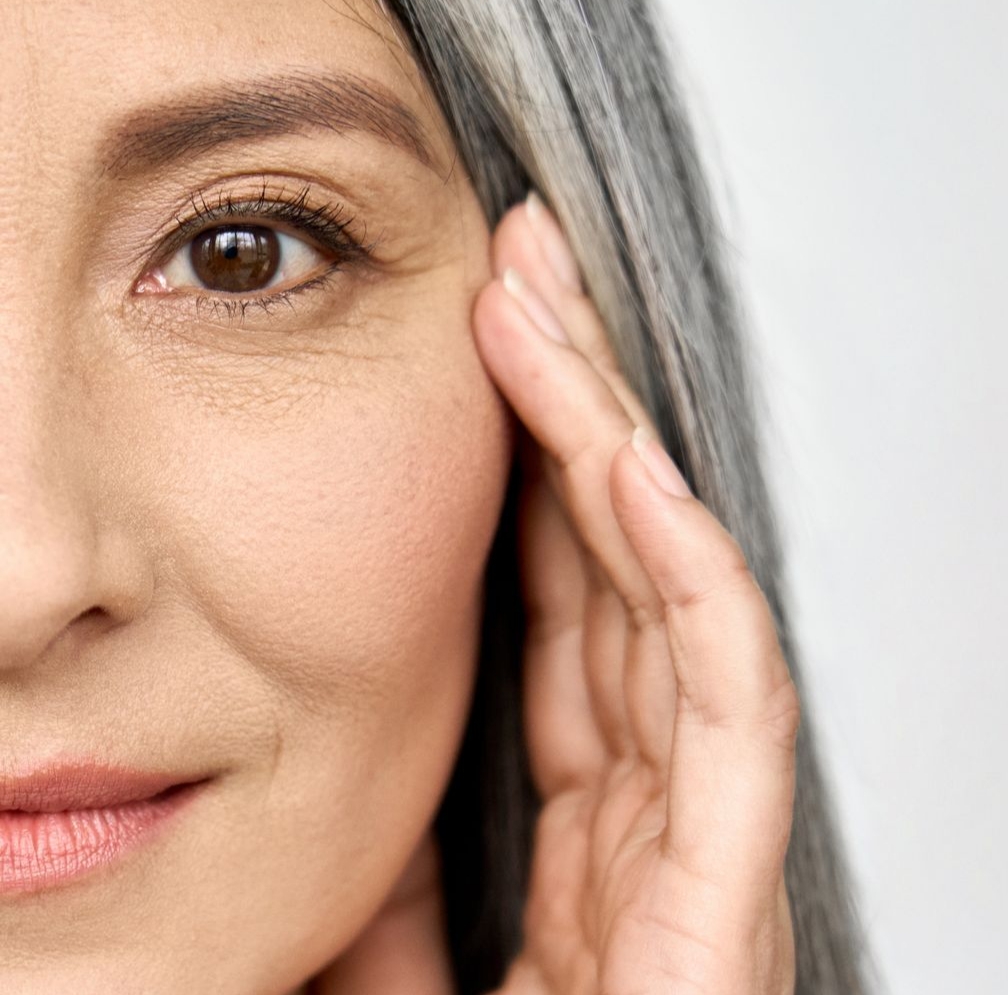 Femme aux cheveux gris, la main près du visage, laissant apparaître les rides autour des yeux.