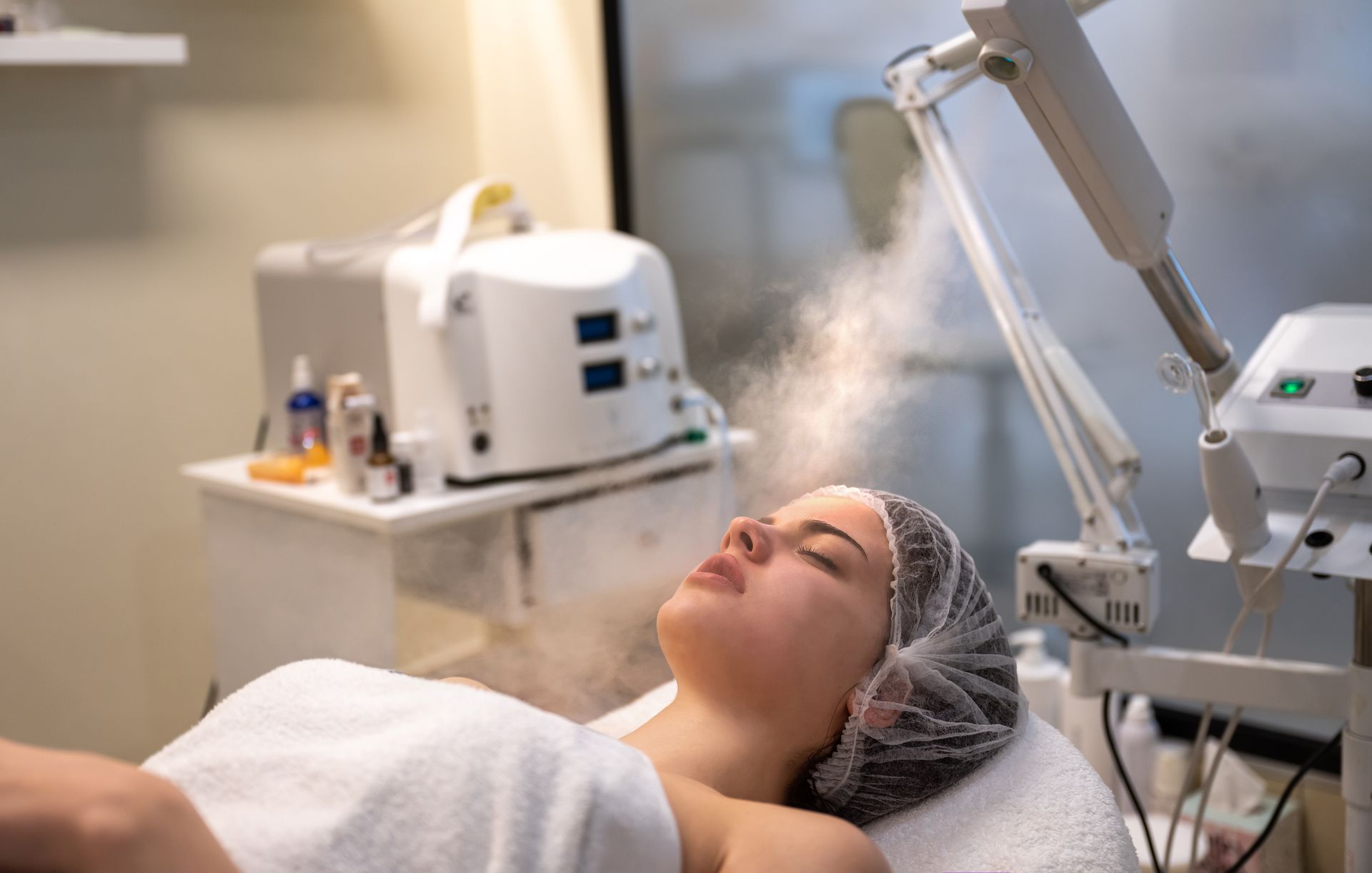 Femme recevant un soin du visage à la vapeur dans un spa, yeux fermés, serviette blanche.