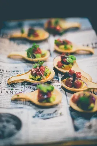 Aperitivos en cucharas de madera, dispuestos sobre papel de periódico antiguo. Cada cuchara contiene comida cortada en cubos y cubierta con microvegetales.