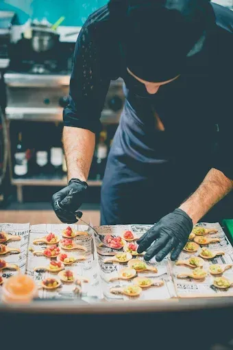 Chef con guantes negros organizando aperitivos en bandejas en una cocina.