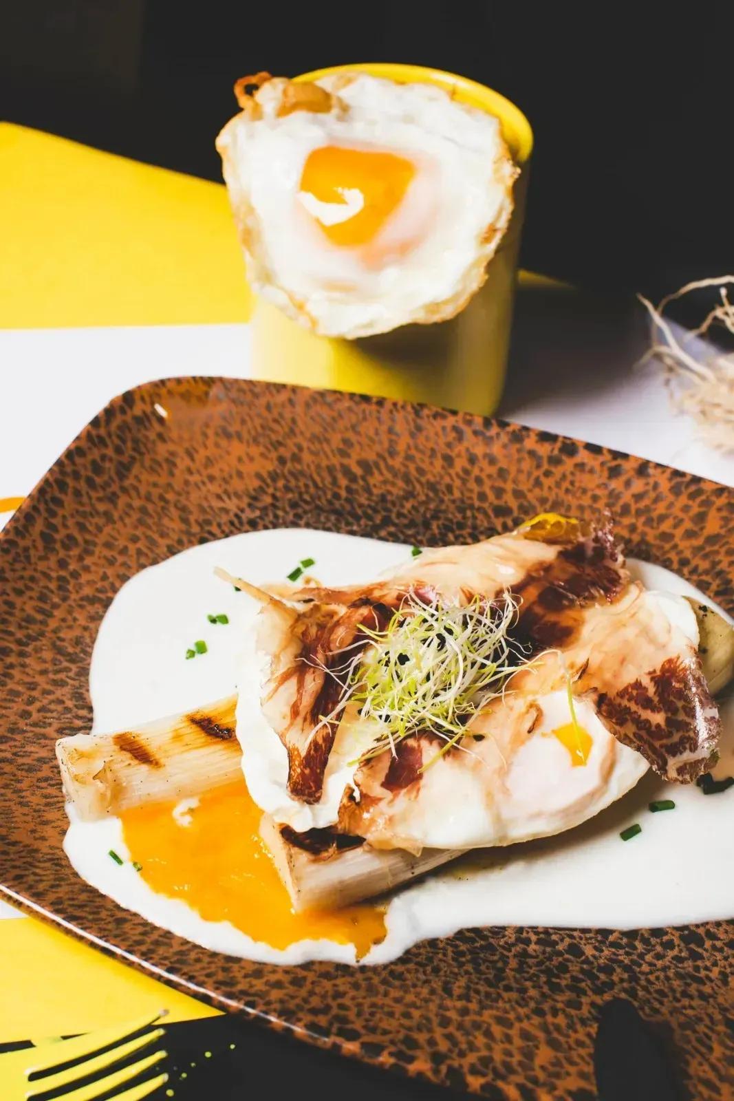Plato de comida con huevo, salsa y espárragos, con un huevo frito en una taza amarilla en el fondo.