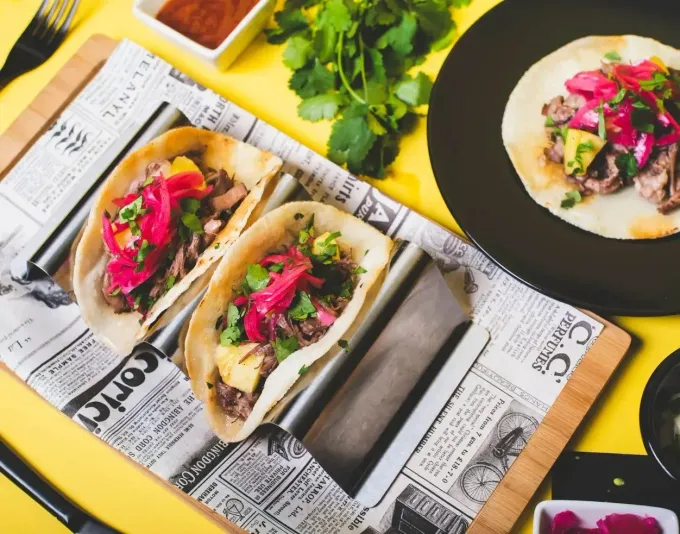 Tacos en una bandeja forrada con papel de periódico, cubiertos con cebollas encurtidas y cilantro, sobre una superficie amarilla.