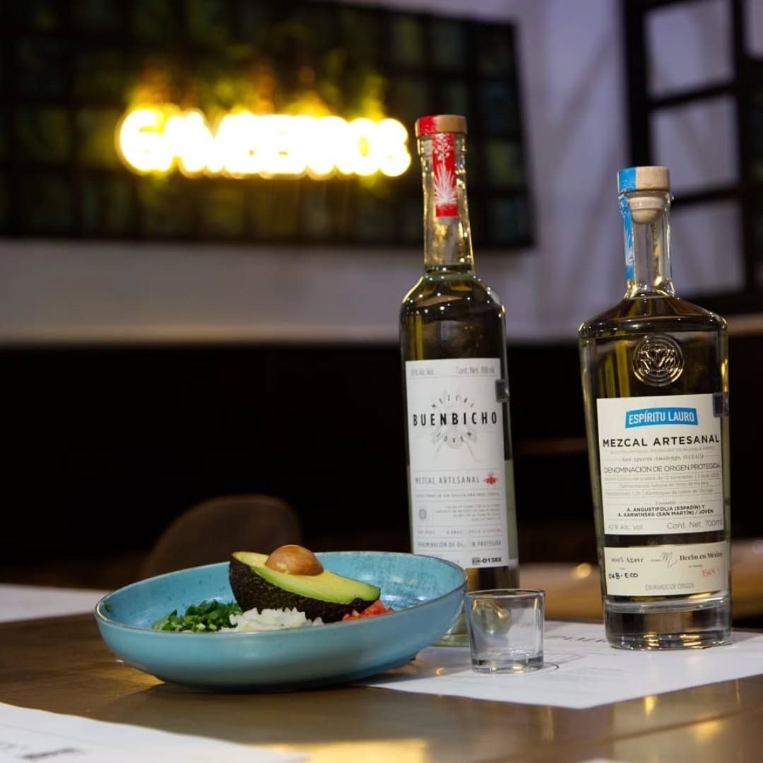 Dos botellas de mezcal, un platito con aguacate y cebolla y un vaso de shot sobre una mesa.