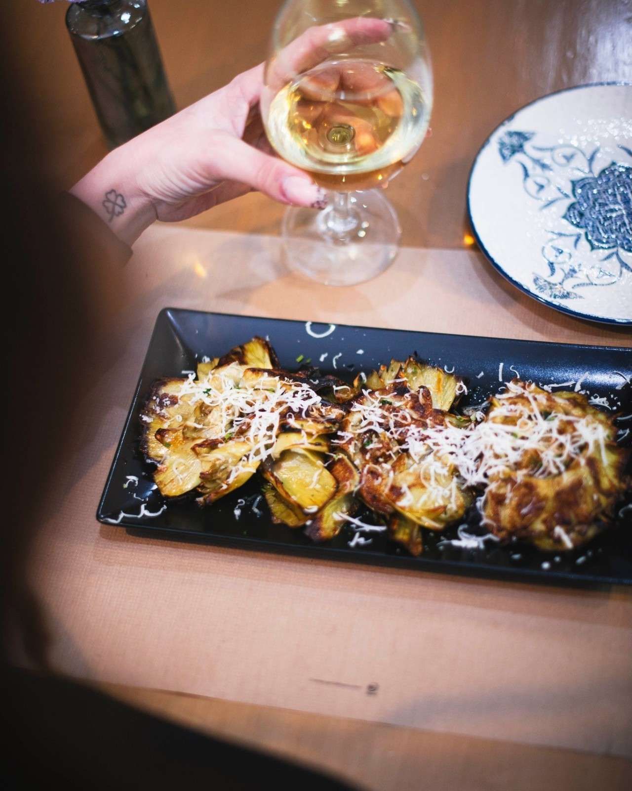 Persona que sostiene una copa de vino y se acerca a un plato de patatas asadas cubiertas con queso.