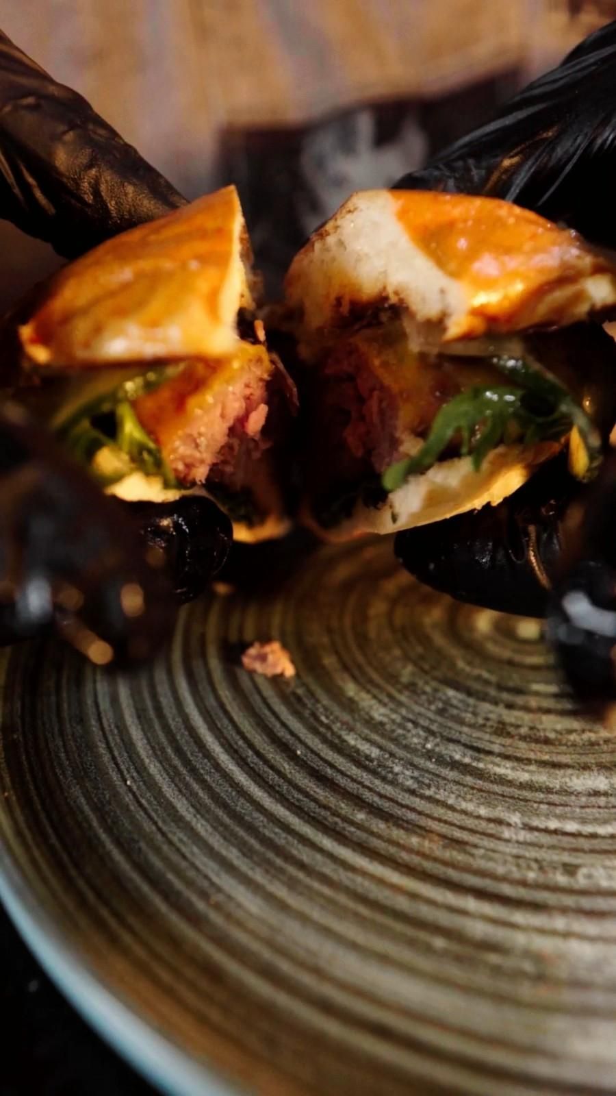 Un sándwich cortado por la mitad, dejando al descubierto el relleno y sostenido por manos enguantadas, reposa sobre un plato estampado.