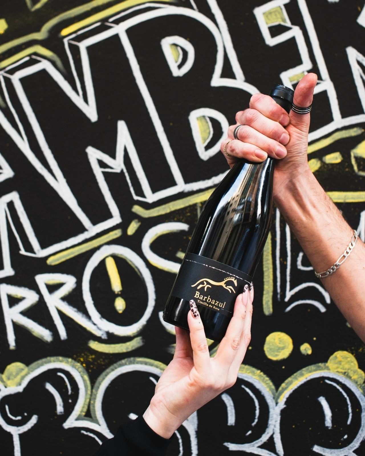 Persona sosteniendo una botella de vino negro con etiqueta dorada, frente a una pared de graffitis.