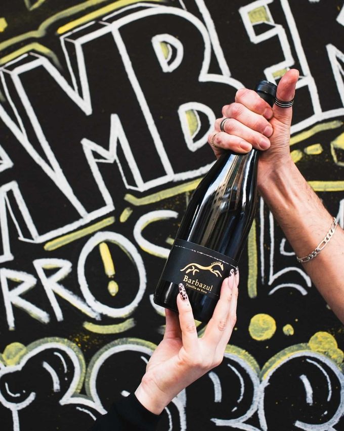Persona sosteniendo una botella de vino negro con etiqueta dorada, frente a una pared de graffitis.