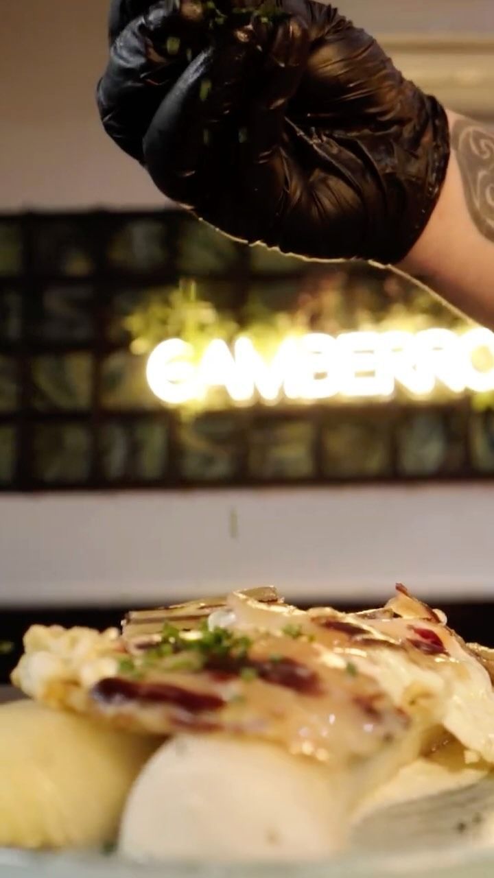 La mano enguantada del chef espolvorea hierbas sobre un plato de pescado con puré de papas, frente a un letrero de neón.