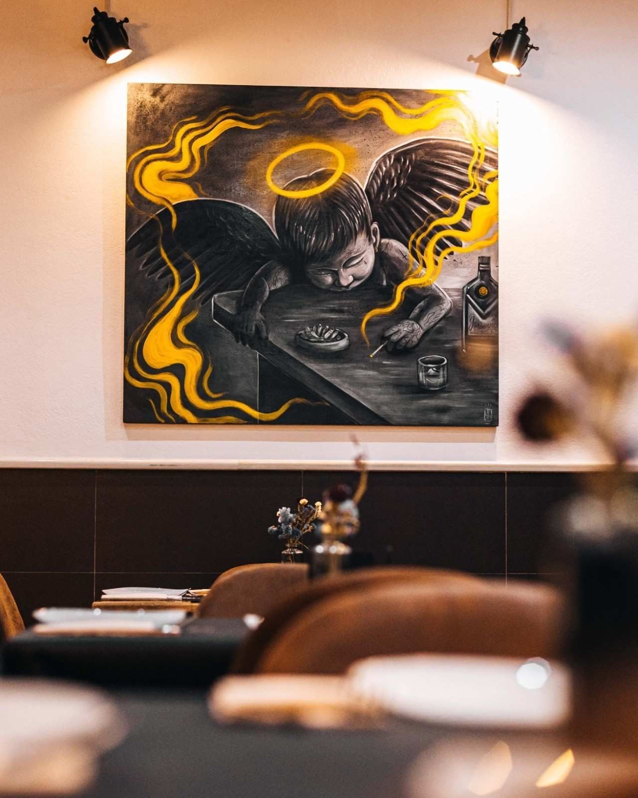 Pintura de un ángel con halo, alas y un cigarrillo sentado a una mesa, iluminado por focos, en un restaurante.