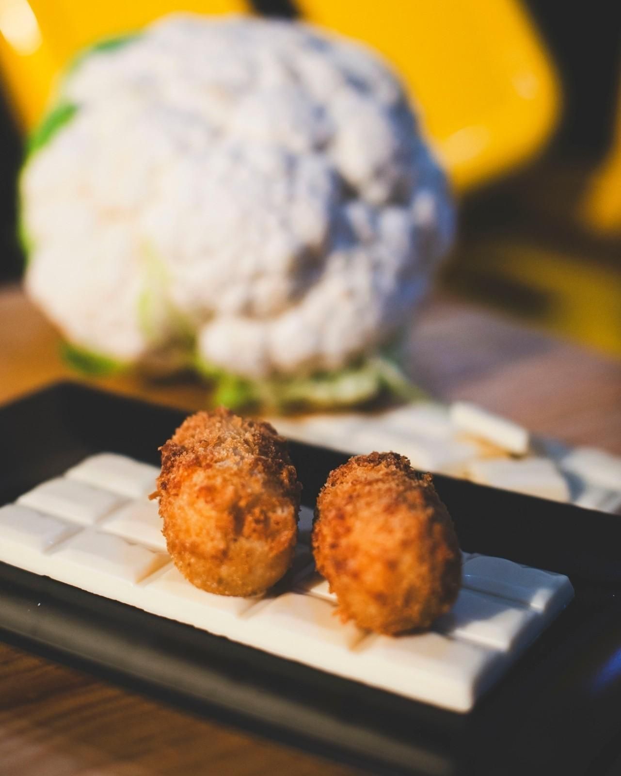 Dos alimentos empanizados y ensartados sobre un bloque blanco, con coliflor en el fondo.
