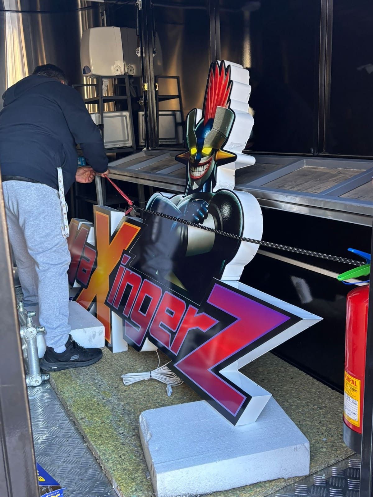 Una persona coloca un gran cartel estilizado del anime Mazinger Z dentro de un vehículo, en el que aparece un robot negro con una cresta roja.