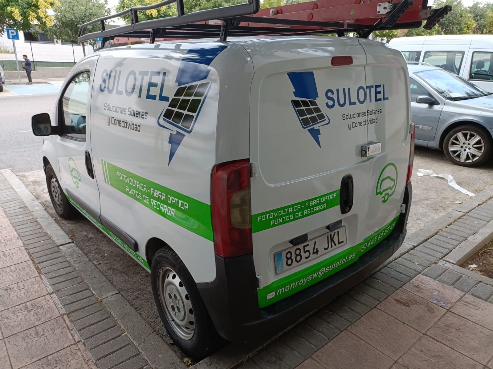 Una furgoneta blanca de la empresa SULOTEL, con la marca en color verde y una baca en el techo, aparcada en una calle asfaltada.