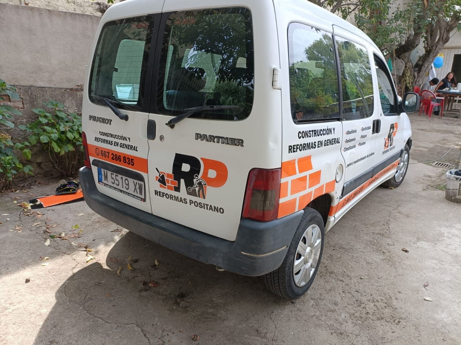 Una furgoneta de trabajo Peugeot Partner blanca con adhesivos de color naranja con estampado de ladrillos, aparcada en un terreno pavimentado.