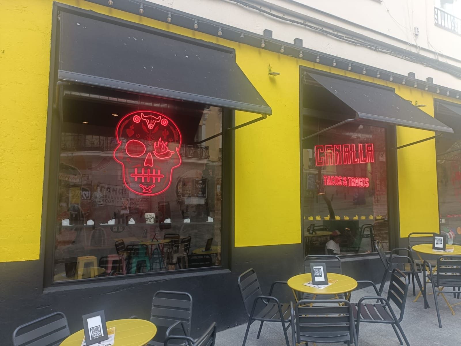 Una fachada amarilla y negra con mesas al aire libre y un letrero de neón rojo con forma de calavera en el escaparate.