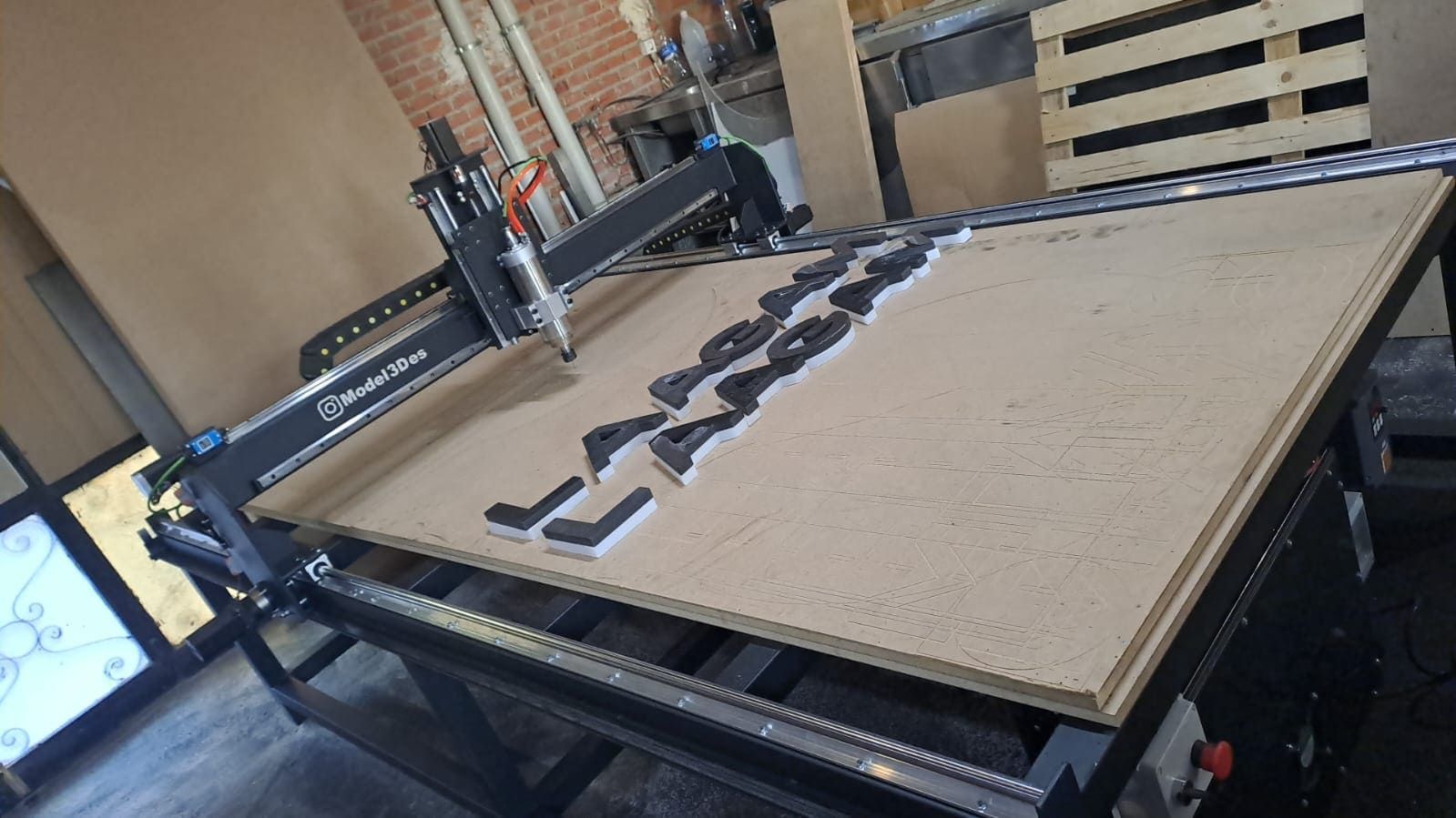Una fresadora CNC corta formas de letras mayúsculas negras a partir de una lámina de madera marrón claro sobre una mesa de taller metálica.