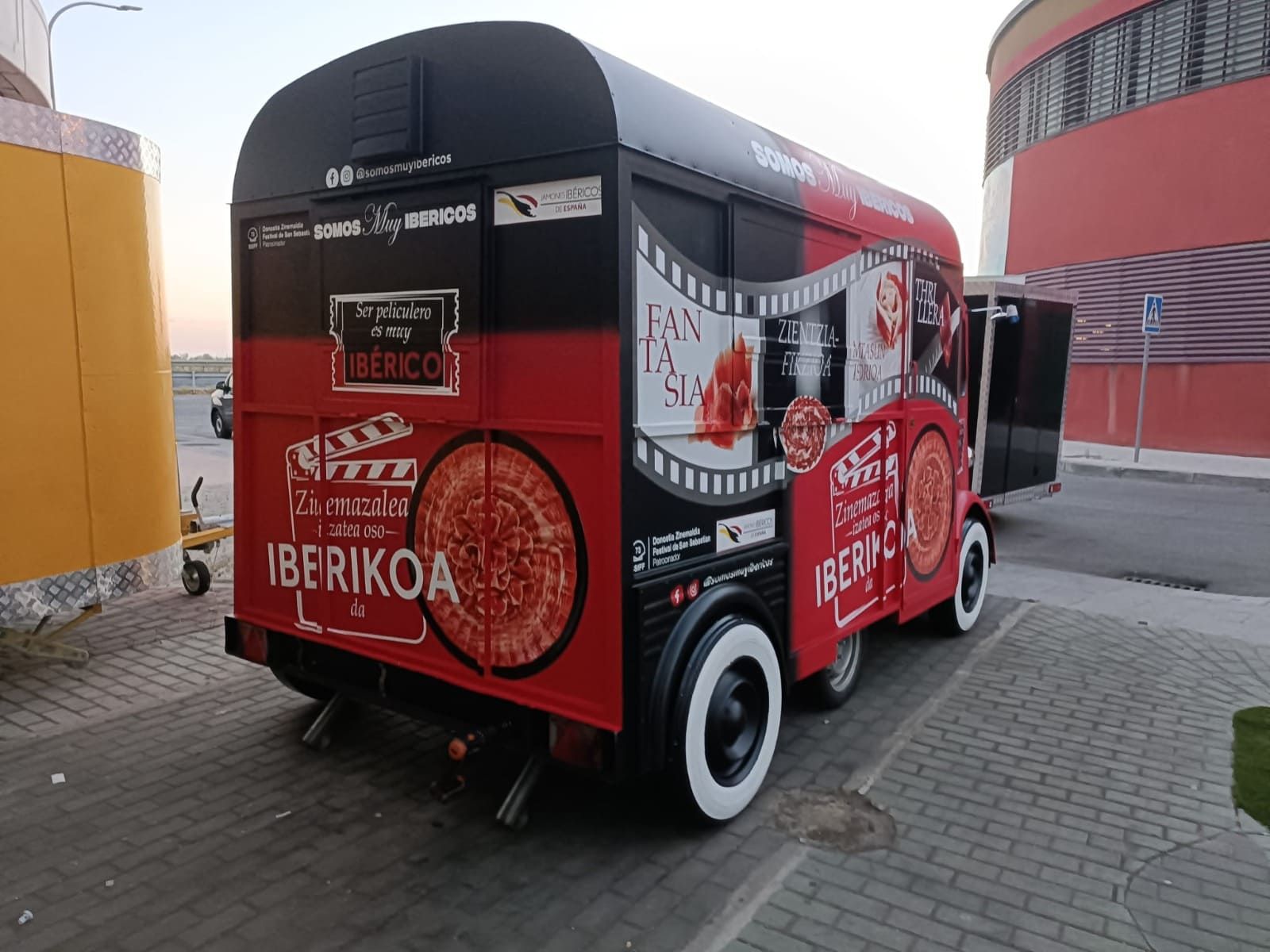 Un camión de comida rojo y negro con la marca "UBERIKOA" y con dibujos de pizza estaba estacionado en una acera de ladrillos junto a un edificio.